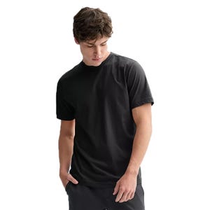A man wearing a plain black T-shirt with short sleeves. He is casually posed with one hand in a trouser pocket, looking downward.
