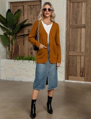A mustard yellow button-up cardigan with two front pockets is worn over a white top, paired with a knee-length denim skirt and black ankle boots.