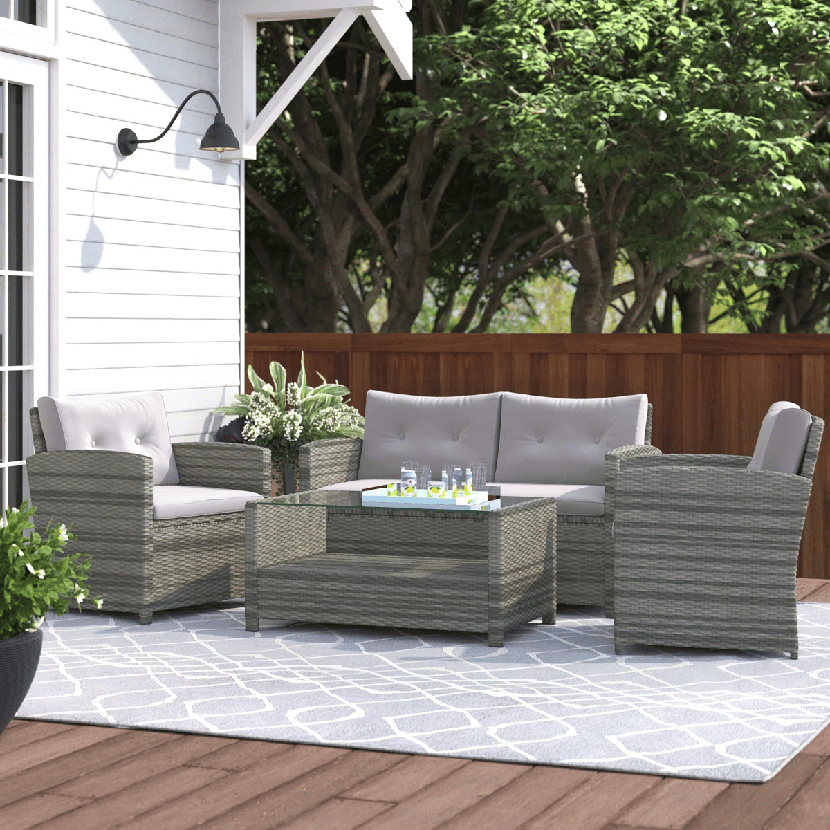 Outdoor patio set with two armchairs, a loveseat, and a glass-top coffee table made from gray wicker, featuring white cushions, set on a patterned rug near a white house wall and wooden fence. Drinks and a plant are placed on the table and beside the seating.