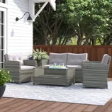 Outdoor patio set with two armchairs, a loveseat, and a glass-top coffee table made from gray wicker, featuring white cushions, set on a patterned rug near a white house wall and wooden fence. Drinks and a plant are placed on the table and beside the seating.