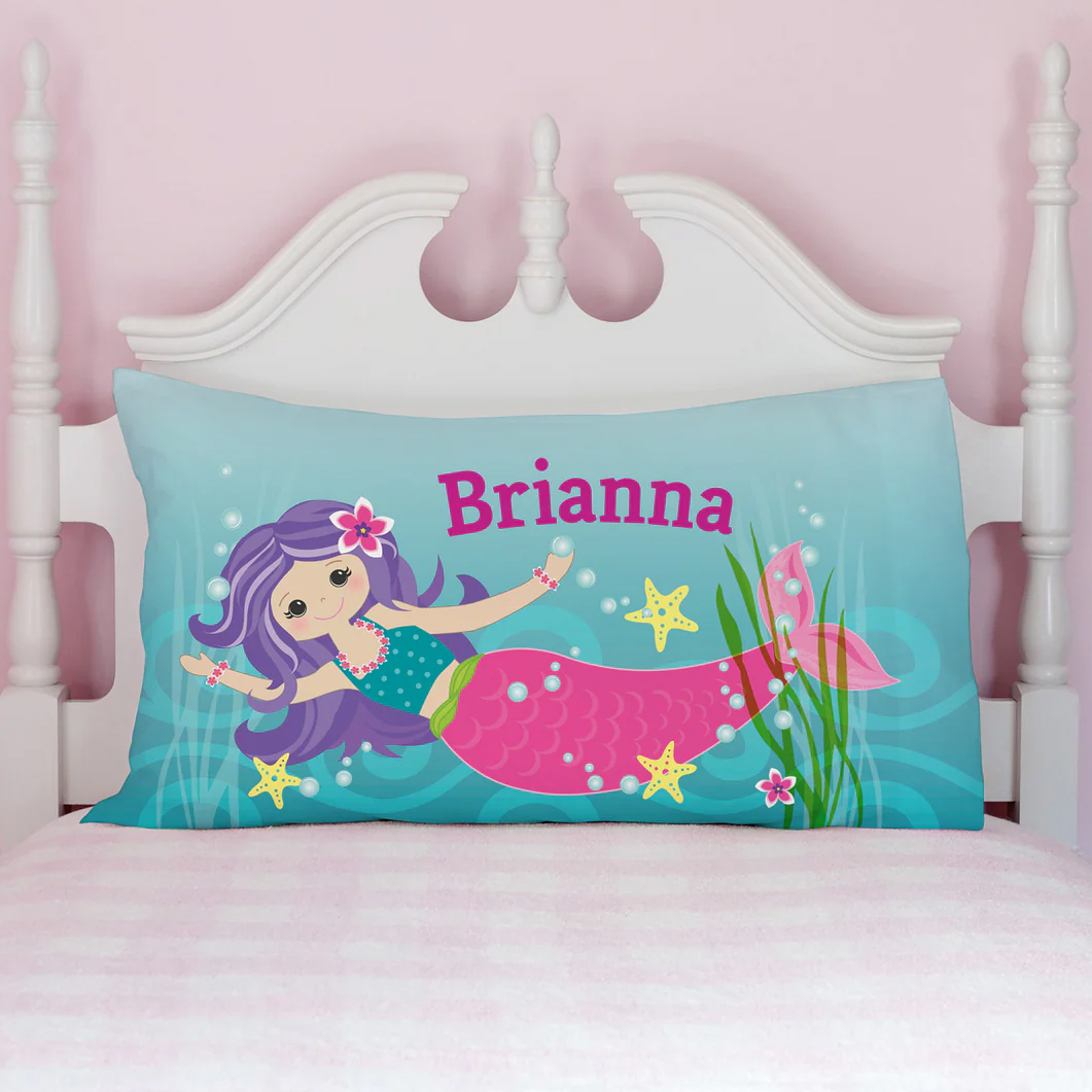 A personalized pillowcase featuring a colorful mermaid design on a teal background with bubbles and starfish, with the name \“Brianna\“ printed in bold pink letters. It's placed on a white headboard and a light pink-striped bedding.