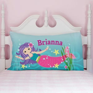 A personalized pillowcase featuring a colorful mermaid design on a teal background with bubbles and starfish, with the name \“Brianna\“ printed in bold pink letters. It's placed on a white headboard and a light pink-striped bedding.