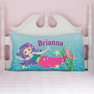 A personalized pillowcase featuring a colorful mermaid design on a teal background with bubbles and starfish, with the name \“Brianna\“ printed in bold pink letters. It's placed on a white headboard and a light pink-striped bedding.