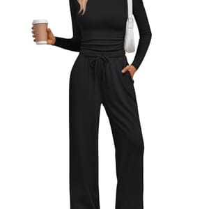 A person wearing a black, long-sleeve, drawstring loungewear set with wide-leg pants and white sneakers, holding a coffee cup and carrying a white shoulder bag.
