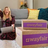 A woman holds a laptop, sitting next to Wayfair boxes on the floor.