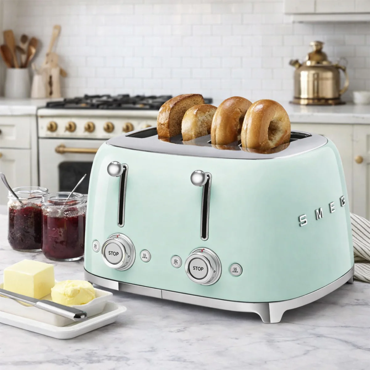 A mint green SMEG 4-slice toaster with toasted bagels is on a kitchen countertop, surrounded by jars of jam and a dish with butter.