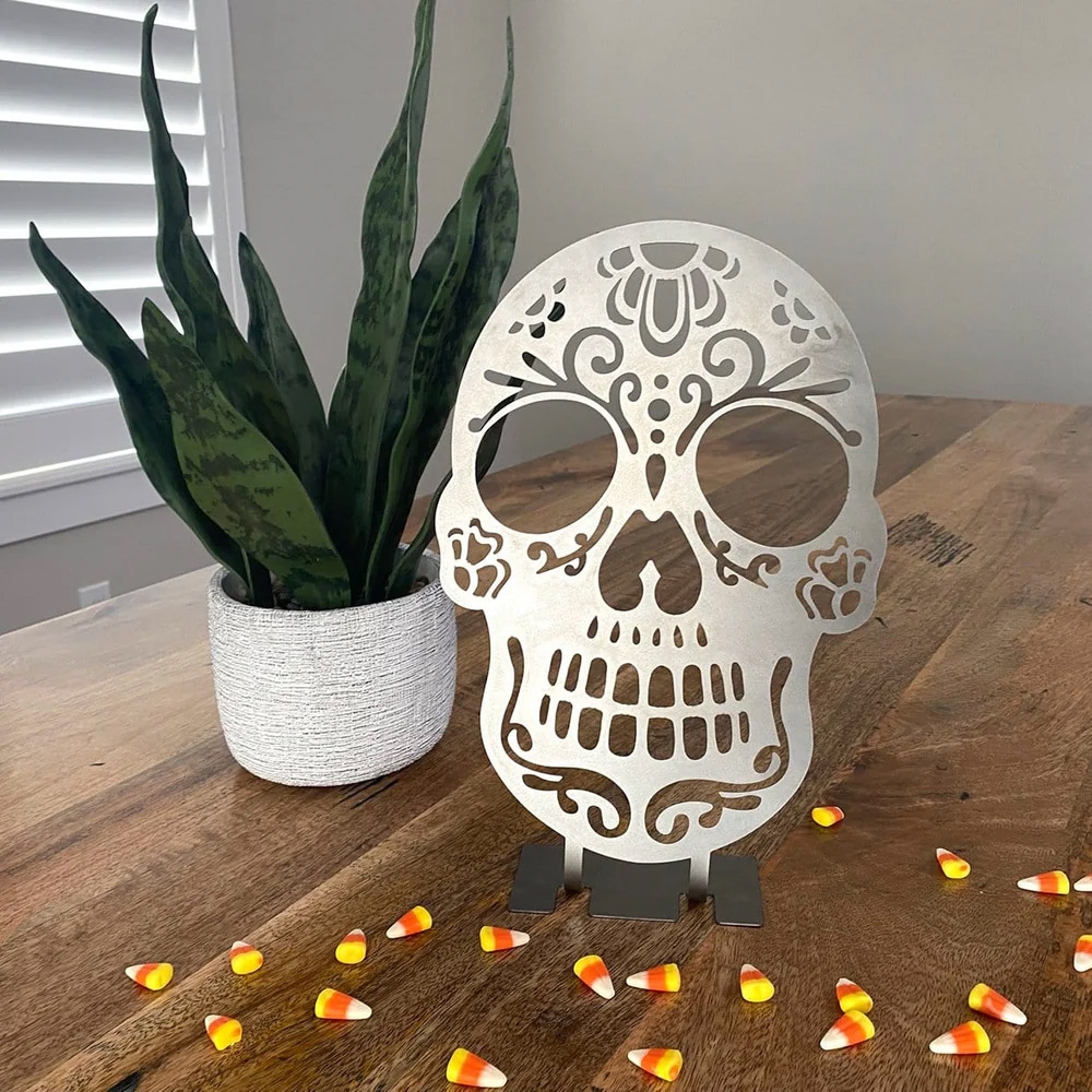 A decorative metal cut-out skull with floral and heart patterns, displayed on a tabletop surrounded by candy corn, adjacent to a potted plant.