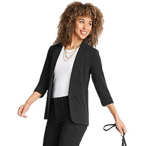 A woman wears a black blazer with three-quarter sleeves over a white top, paired with black pants. She holds a black strap and wears layered necklaces.