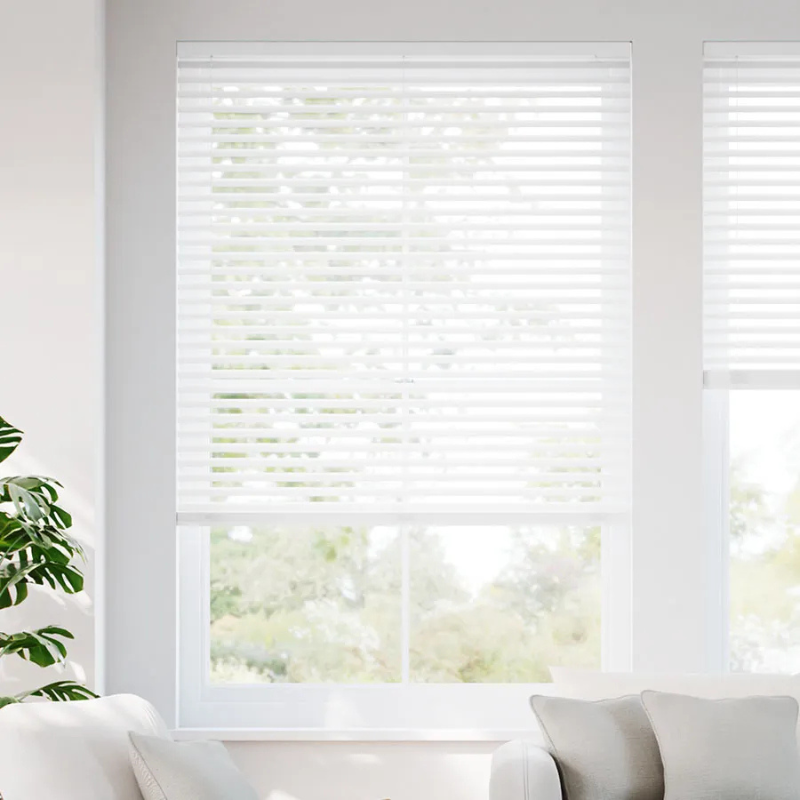 White horizontal blinds partially lowered on a window with a view of greenery outside; a partial corner of a light-colored sofa and a green plant are visible inside.