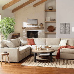 A living room setup with sectional sofa, round coffee table, side stool, area rug, decorative wall art, floating shelves, and indoor plants.