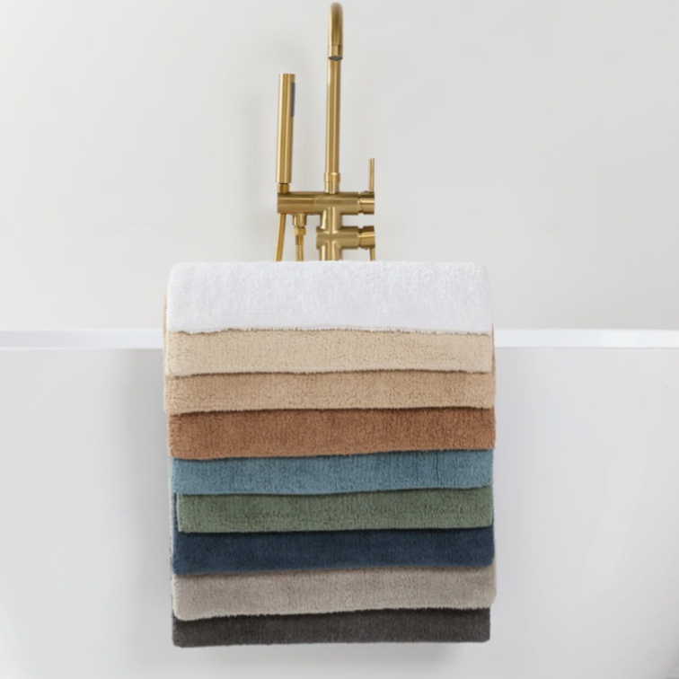 A selection of neatly stacked towels in various colors is draped over the side of a modern white bathtub with a gold faucet, set on a marble-tiled floor.
