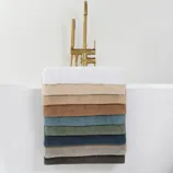 A selection of neatly stacked towels in various colors is draped over the side of a modern white bathtub with a gold faucet, set on a marble-tiled floor.