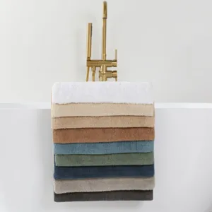 A selection of neatly stacked towels in various colors is draped over the side of a modern white bathtub with a gold faucet, set on a marble-tiled floor.