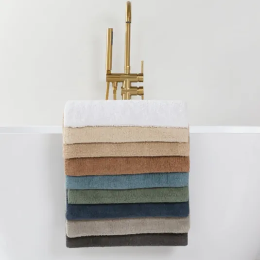 A selection of neatly stacked towels in various colors is draped over the side of a modern white bathtub with a gold faucet, set on a marble-tiled floor.