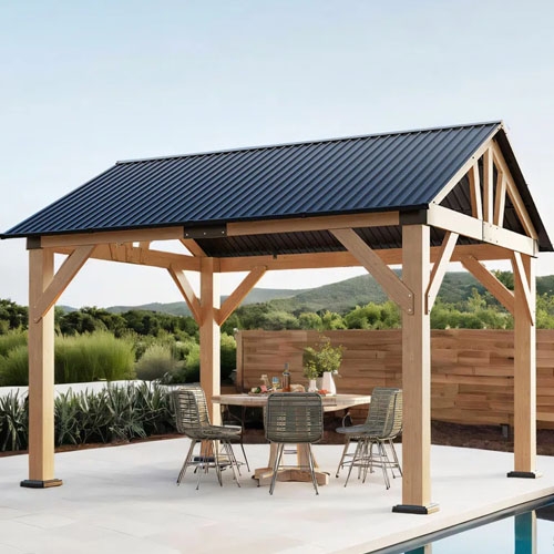 A wooden gazebo with a dark metal roof stands on a patio, featuring a round table and four wicker chairs. The backdrop includes greenery and hills.