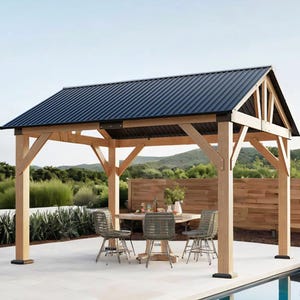 A wooden gazebo with a dark metal roof stands on a patio, featuring a round table and four wicker chairs. The backdrop includes greenery and hills.
