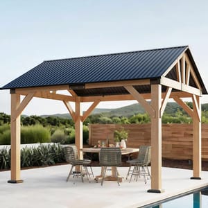 A wooden gazebo with a dark metal roof stands on a patio, featuring a round table and four wicker chairs. The backdrop includes greenery and hills.