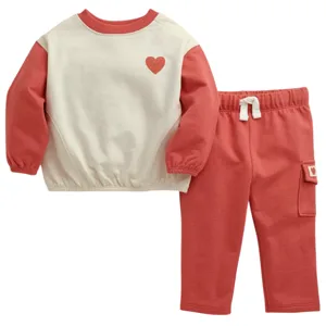A two-piece children's outfit featuring a cream and red long-sleeve sweatshirt with a small heart design, paired with matching red pants with a tie waistband and a pocket.