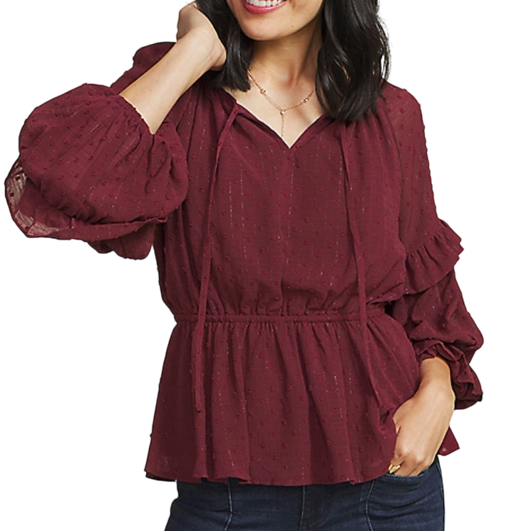 A woman is wearing a maroon blouse with textured dot patterns, featuring long balloon sleeves, a V-neck with ties, and a peplum waist. She's paired it with dark jeans and a delicate necklace.
