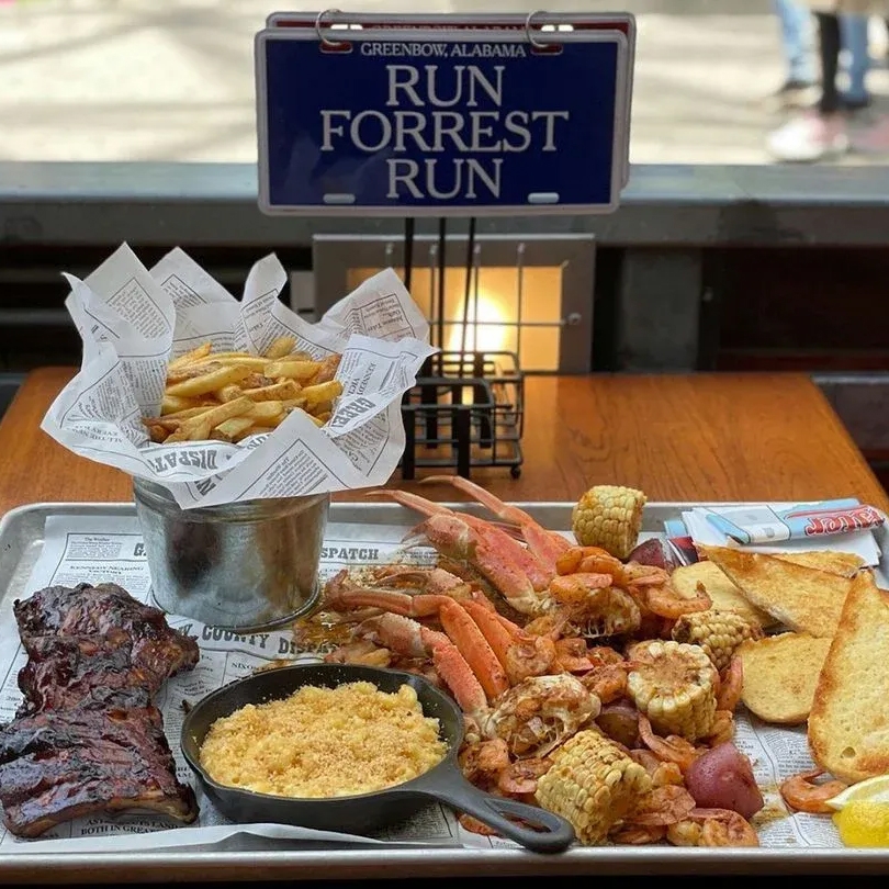 A tray features a seafood boil with crab, shrimp, corn, potatoes, and toast, accompanied by ribs, fries in a metal bucket, and mac and cheese in a skillet. A sign reads \“RUN FORREST RUN\“ in the background.
