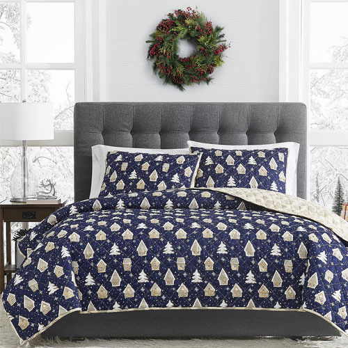3pc Reversible Gingerbread Quilt Set features a navy quilt and pillow shams with beige gingerbread house and tree designs, reversible to a light beige snowflake pattern, complemented by a gray tufted headboard and festive wreath above.