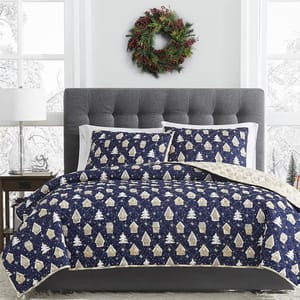 3pc Reversible Gingerbread Quilt Set features a navy quilt and pillow shams with beige gingerbread house and tree designs, reversible to a light beige snowflake pattern, complemented by a gray tufted headboard and festive wreath above.