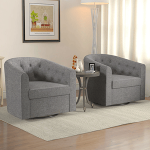 Two gray swivel accent chairs with a tufted design and curved armrests are set on a beige patterned rug. They are positioned around a small round side table with decorative items, under a framed wall picture and next to a floor lamp.