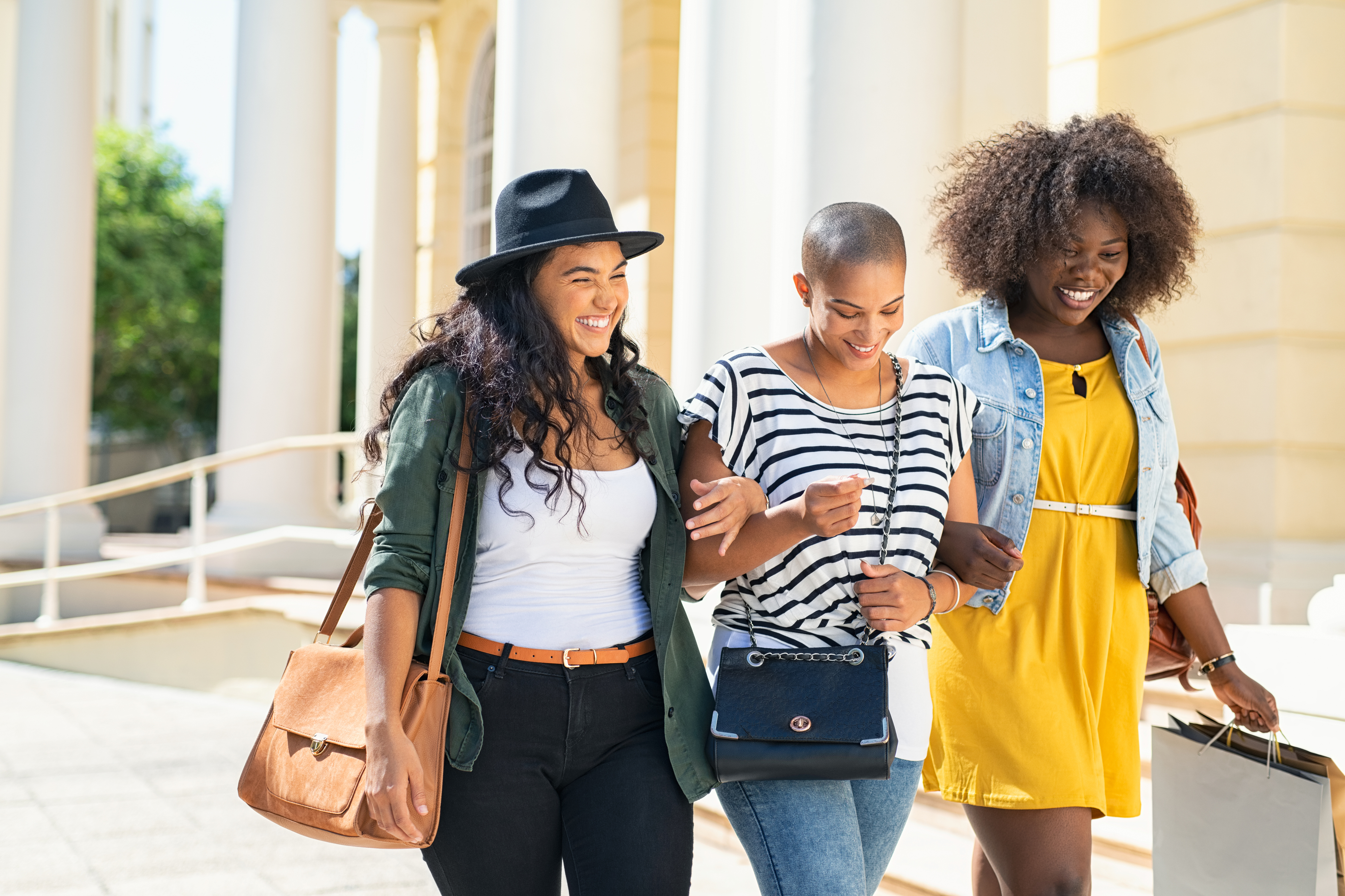 Three women walking together, one with a brown shoulder bag, another with a black crossbody bag, and the third carrying shopping bags.