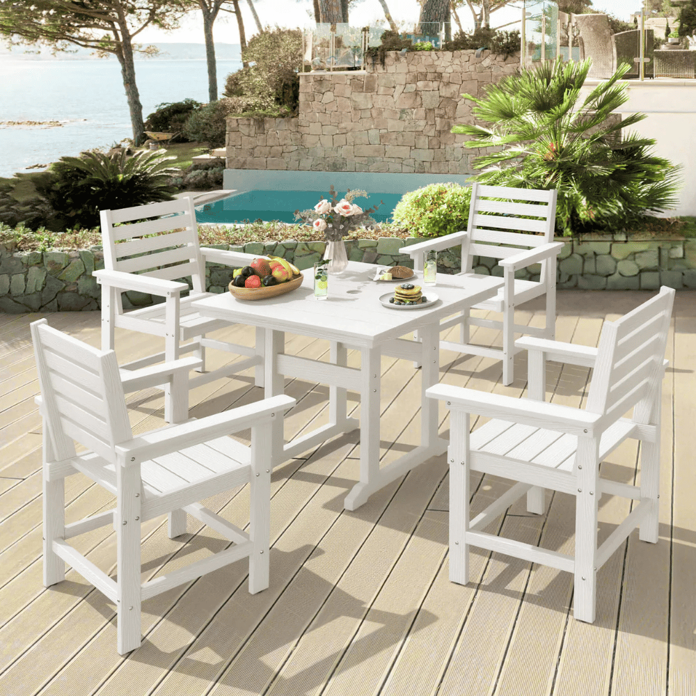 White outdoor dining set with four chairs and a table on a deck, featuring a fruit bowl, drinks, and flowers. The setting overlooks a swimming pool and scenic water view surrounded by greenery.