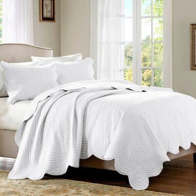 A white quilt set with scalloped edges and textured patterns, including two matching pillow shams, displayed on a bed in a room with natural light.