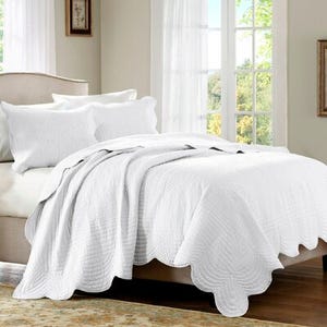 A white quilt set with scalloped edges and textured patterns, including two matching pillow shams, displayed on a bed in a room with natural light.