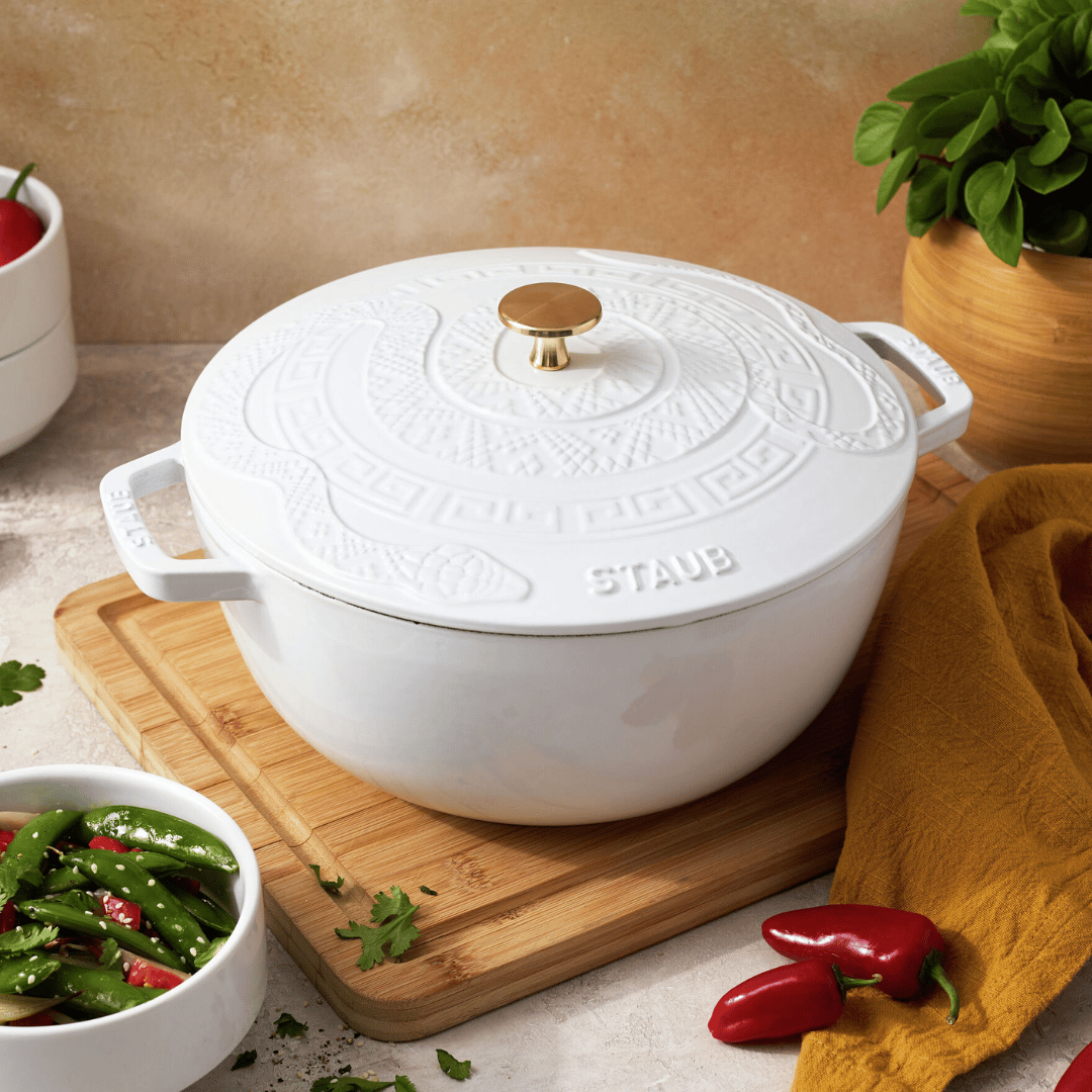 White Staub cast iron cocotte with embossed design and gold knob, placed on a wooden board, surrounded by a bowl of vegetables and fresh herbs.