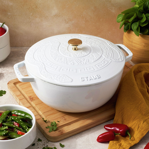 White Staub cast iron cocotte with embossed design and gold knob, placed on a wooden board, surrounded by a bowl of vegetables and fresh herbs.