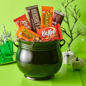 A cauldron filled with an assortment of candy bars, including Reese's, Kit Kat, and Hershey's, set against a green background with Halloween decor.