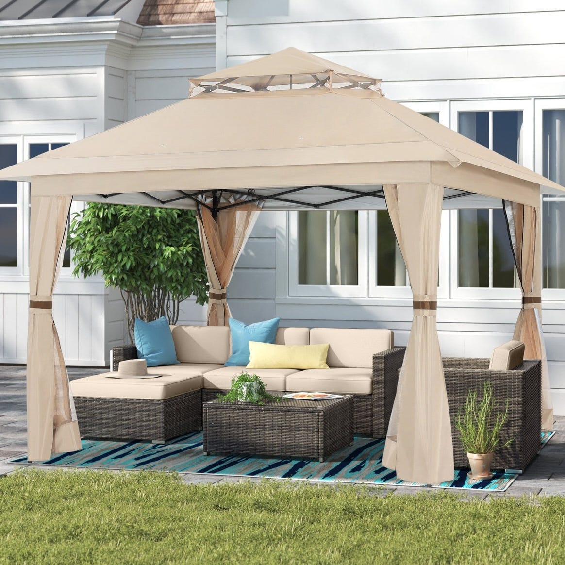 Outdoor patio setup with a beige canopy, wicker sectional sofa, and a coffee table on a blue patterned rug. The setup includes light-colored cushions, two decorative pillows, a small potted plant, and a privacy curtain.