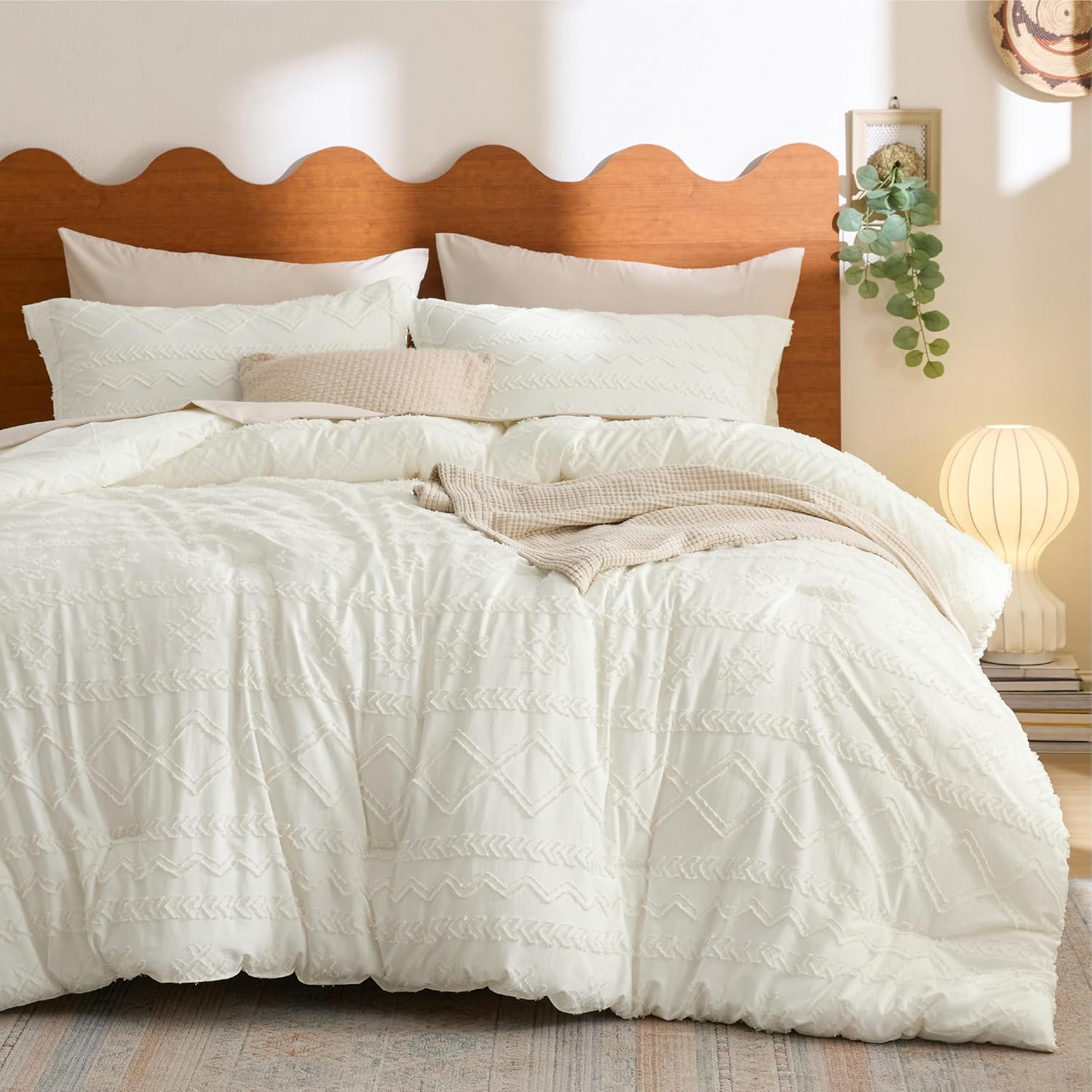 A bed features a white textured comforter with geometric patterns, matching pillows, a wooden scalloped headboard, a small throw blanket, and a round table lamp on stacked books.