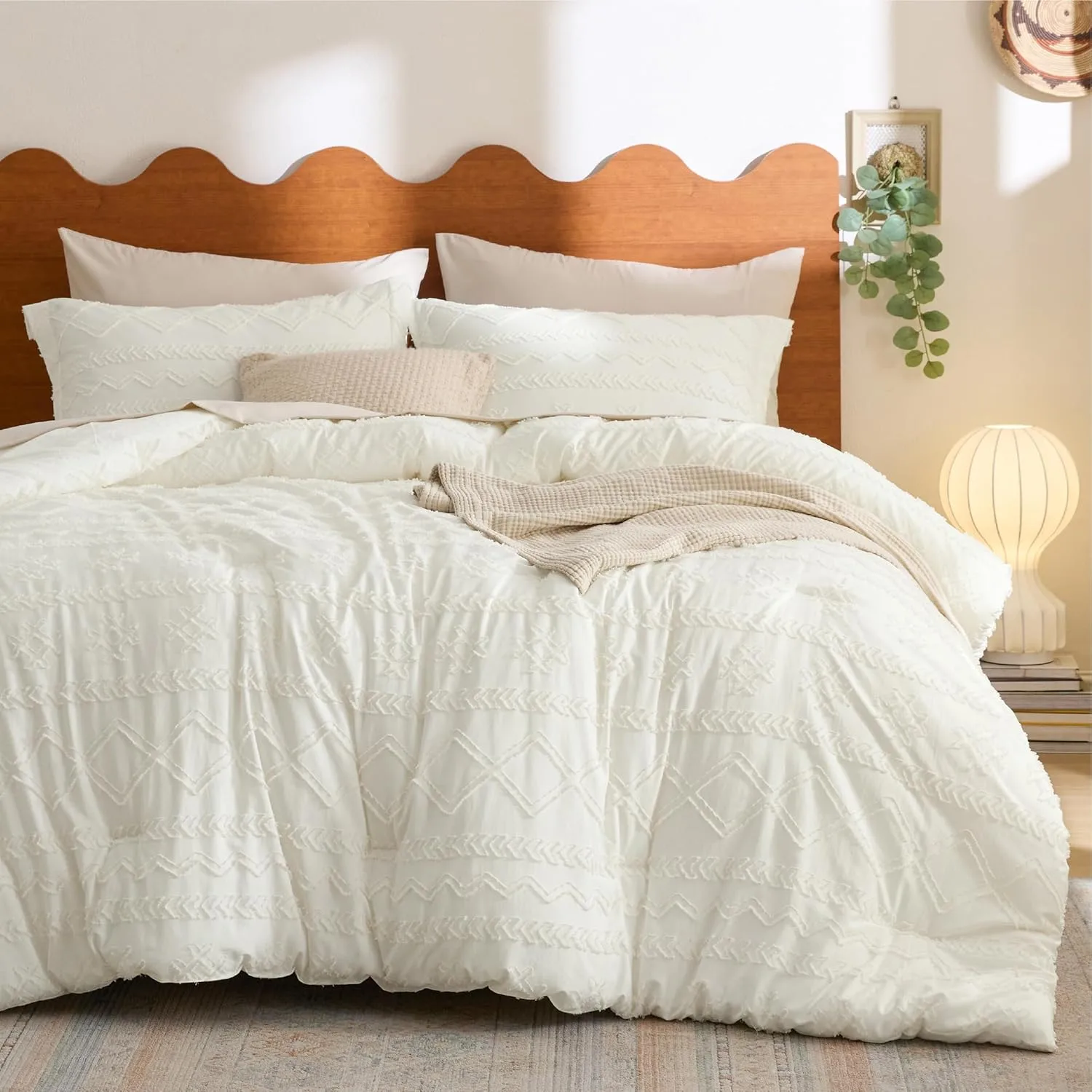 A bed features a white textured comforter with geometric patterns, matching pillows, a wooden scalloped headboard, a small throw blanket, and a round table lamp on stacked books.