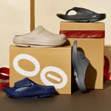Various styles of OOFOS footwear, including beige clogs, black and navy sandals, are displayed on and alongside branded boxes with a glittery red ribbon.