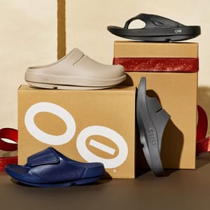 Various styles of OOFOS footwear, including beige clogs, black and navy sandals, are displayed on and alongside branded boxes with a glittery red ribbon.