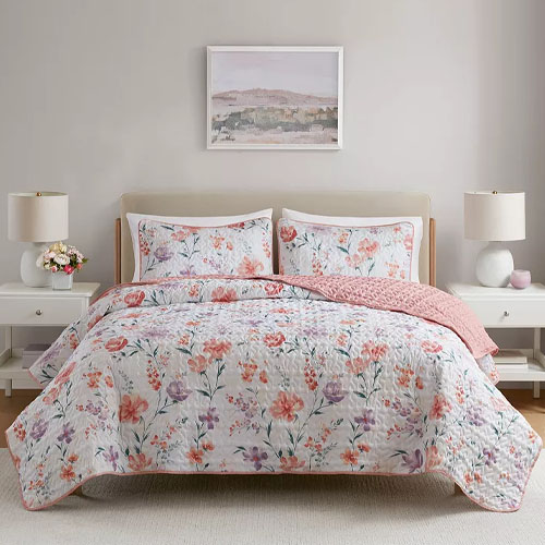 A bed with floral-patterned bedding and two matching pillowcases, complemented by a pink blanket. The setup includes two bedside tables with lamps and decorative items, set against a neutral wall with a landscape painting above the bed.