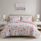 A bed with floral-patterned bedding and two matching pillowcases, complemented by a pink blanket. The setup includes two bedside tables with lamps and decorative items, set against a neutral wall with a landscape painting above the bed.