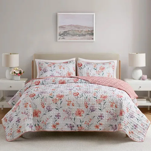 A bed with floral-patterned bedding and two matching pillowcases, complemented by a pink blanket. The setup includes two bedside tables with lamps and decorative items, set against a neutral wall with a landscape painting above the bed.