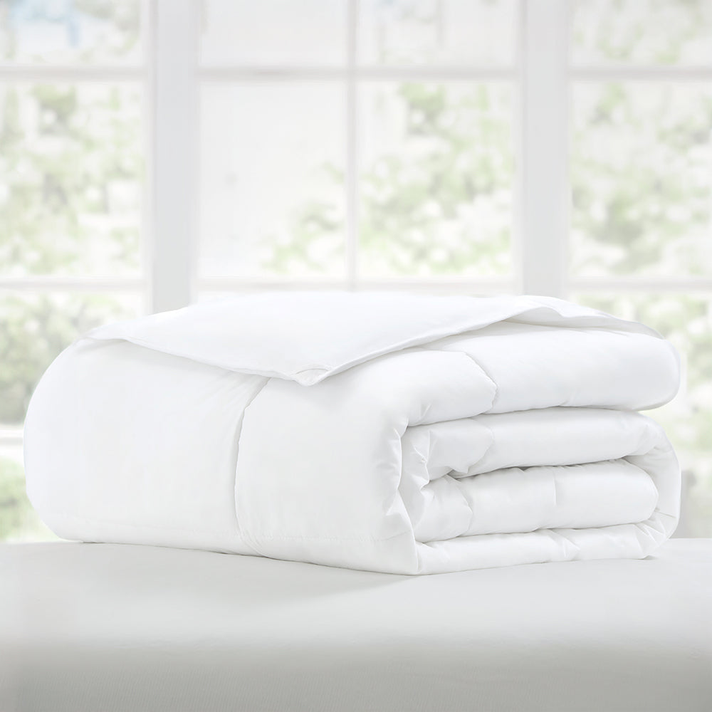 A white comforter is neatly folded on a bed, set against a blurred background of a window with greenery outside.