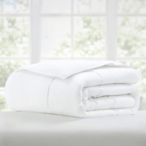 A white comforter is neatly folded on a bed, set against a blurred background of a window with greenery outside.