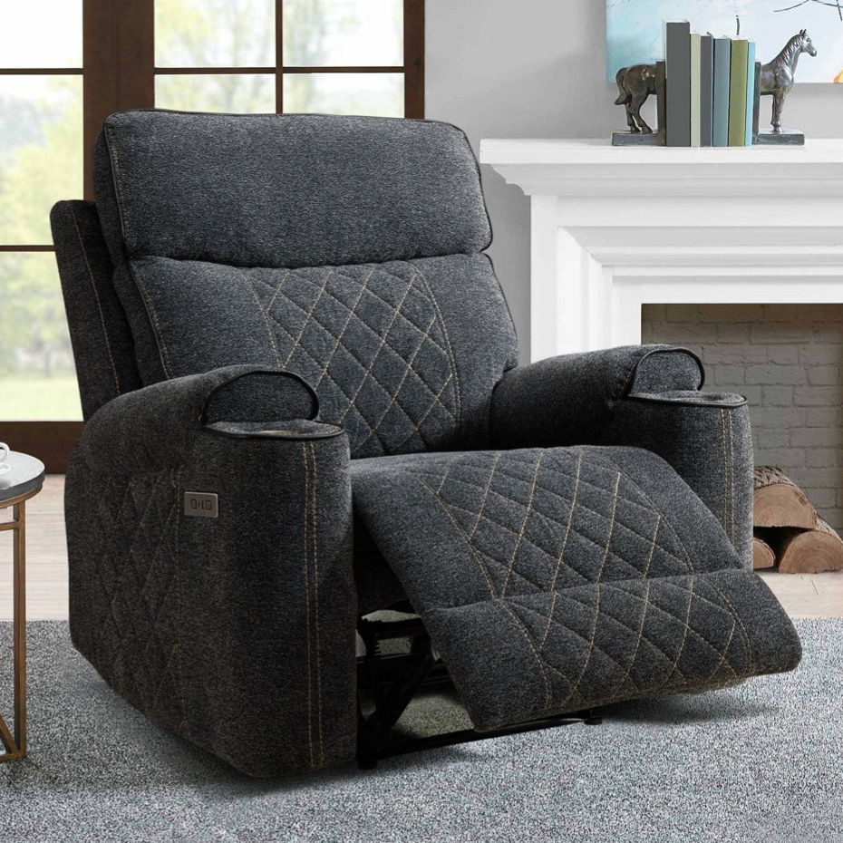 A dark gray, quilted reclining chair with cup holders and control buttons next to a small side table with a lamp and teacups, set in a cozy living room with a fireplace and large window.