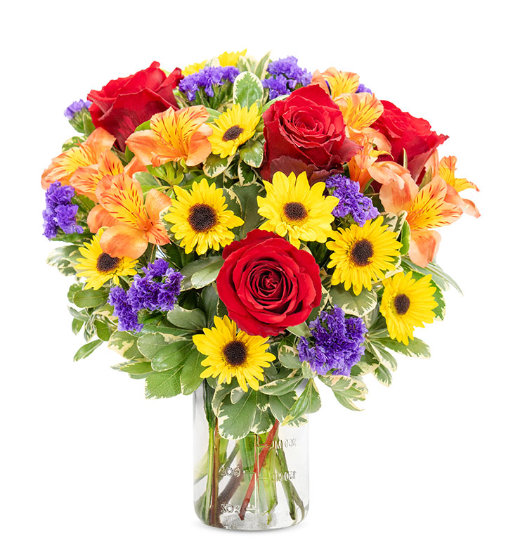 A vibrant bouquet of red roses, yellow sunflowers, orange alstroemeria, and purple statice in a clear glass vase.
