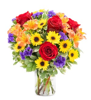 A vibrant bouquet of red roses, yellow sunflowers, orange alstroemeria, and purple statice in a clear glass vase.