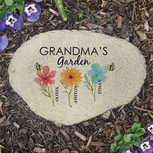 A personalized garden stone features the text \“Grandma's Garden\“ with colorful flowers labeled with the names Olivia, Ashton, and Ethan, set against a natural mulch background with purple and white flowers.