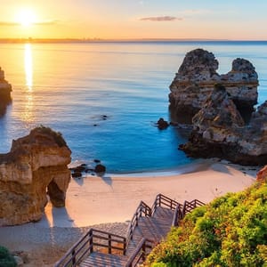 A scenic sunset over a tranquil beach with clear waters, surrounded by unique rock formations and accessed by a wooden staircase.