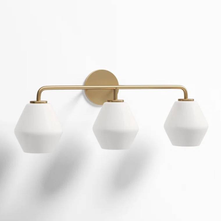 A modern vanity light featuring three geometric white shades with a sleek brass frame, mounted on a circular brass base. It's a dimmable fixture designed for stylish and functional lighting.